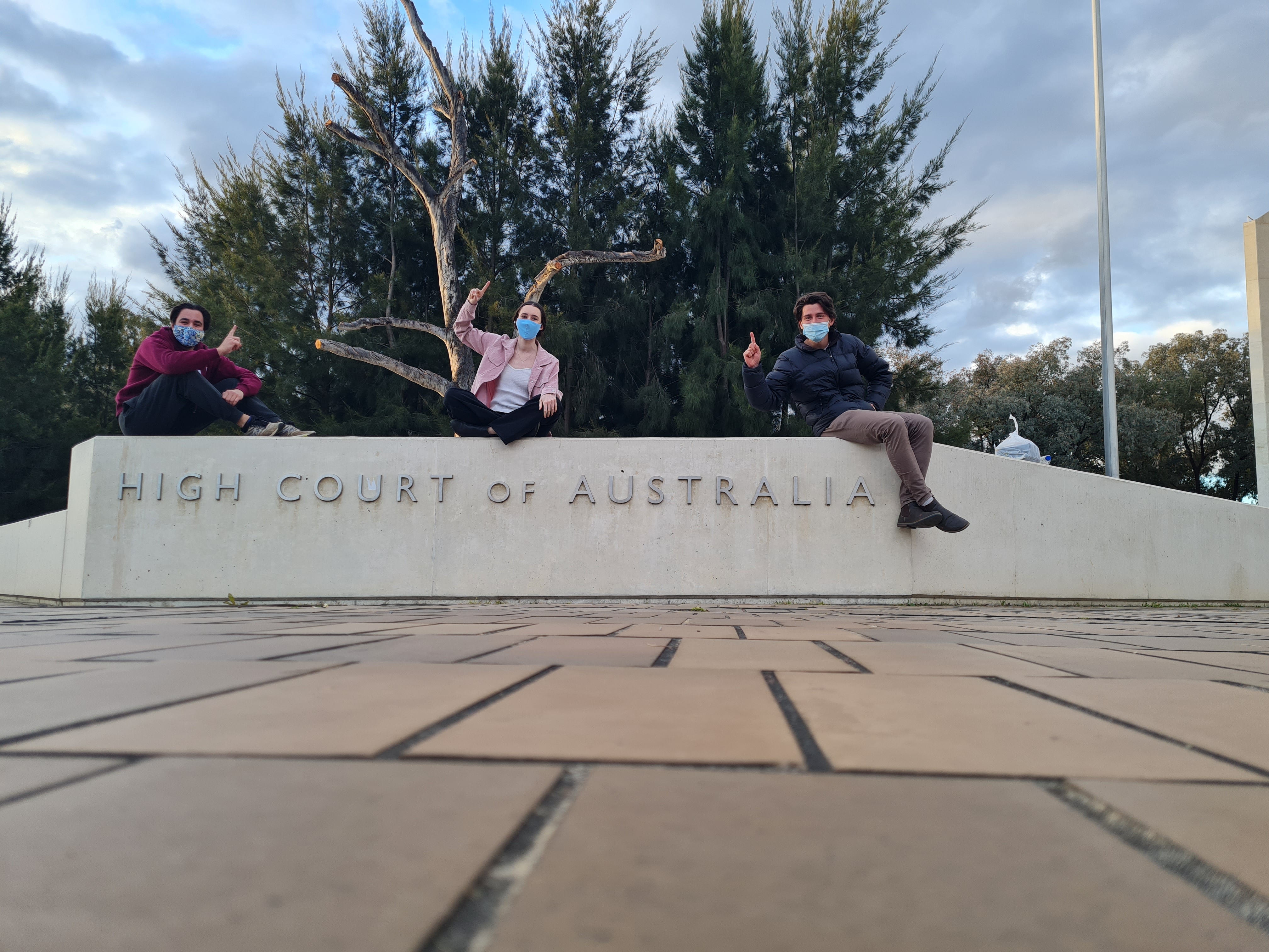 The winning ANU team members Tom Dunbabin, Madeleine McGregor, and Benjamin Durkin, sitting on top of the High Court of Australia sign.