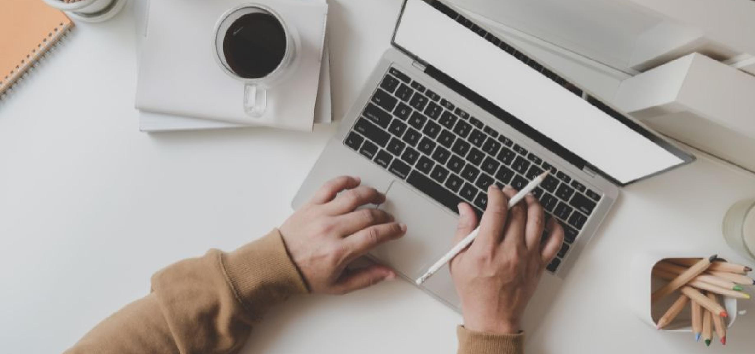 An overhead shot of pair of hands on a laptop next to cups of pencils and coffee